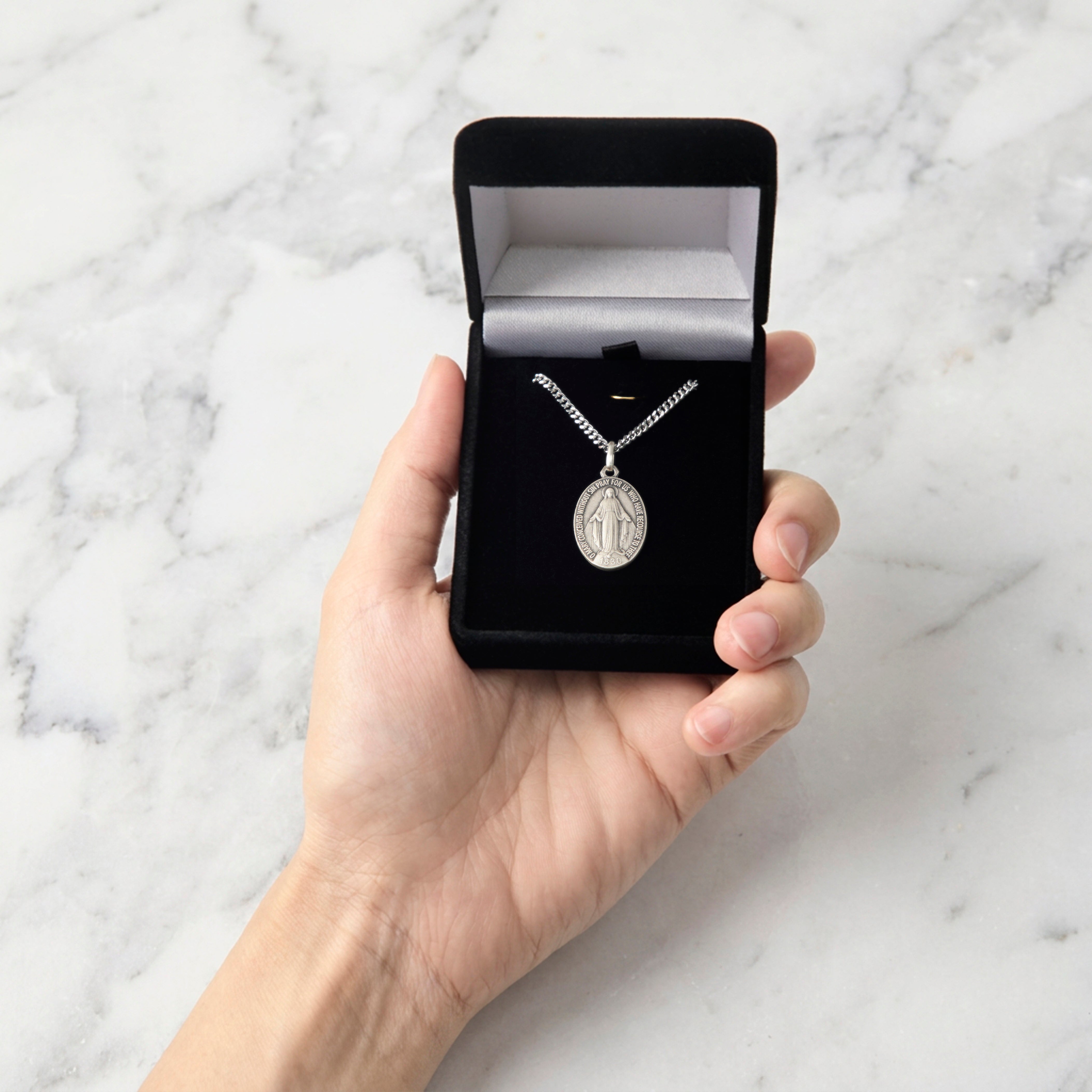 A hand holding an open black jewelry box containing a silver chain necklace with an oval pendant, set against a white marble background.