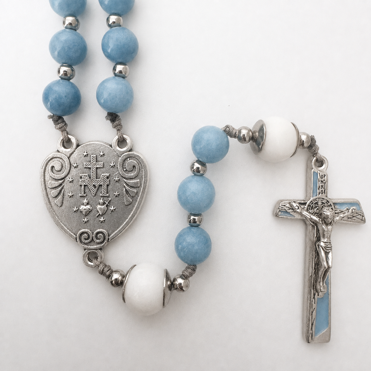 A rosary necklace featuring light blue and white beads, a silver-tone heart-shaped Medallion of Our Lady of Mount Carmel, and a matching crucifix with a light blue enamel inlay, displayed on a clean white background.