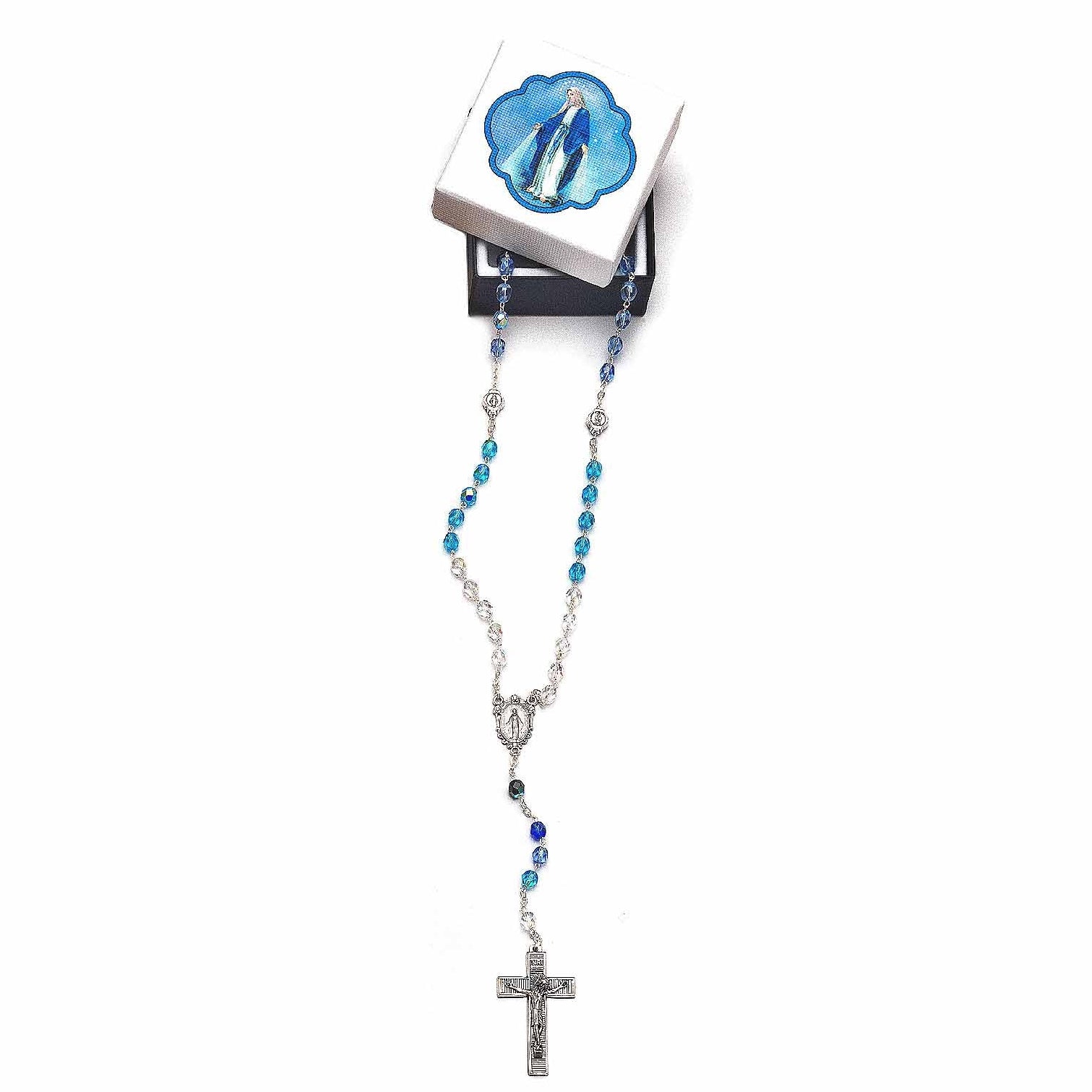 A rosary with blue and silver beads, a silver crucifix pendant, and a small white box with a blue illustration of the Virgin Mary, displayed on a black stand against a white background.
