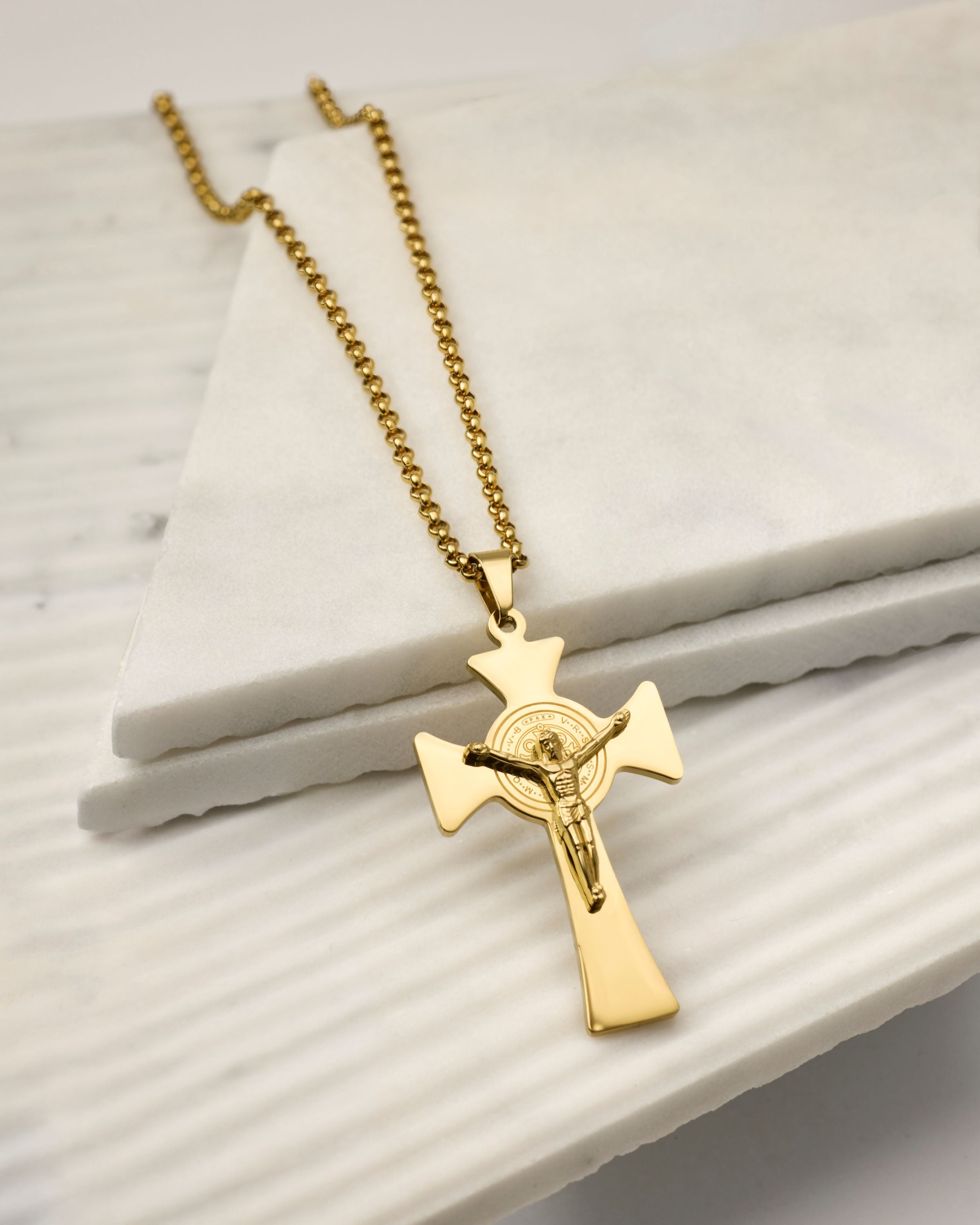 A gold crucifix pendant necklace on a gold ball chain, displayed on stacked white marble blocks.